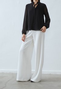 Blusa nera trasparente a maniche lunghe con colletto a fiocco, abbinata a pantaloni bianchi a gamba larga. Tessuto morbido; vestibilità comoda; dettagli minimi.