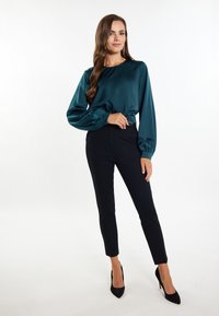 Teal satijnen blouse met lange puffmouwen en geplooide manchetten, gecombineerd met slanke zwarte broek en zwarte stilettos, tegen een witte achtergrond.