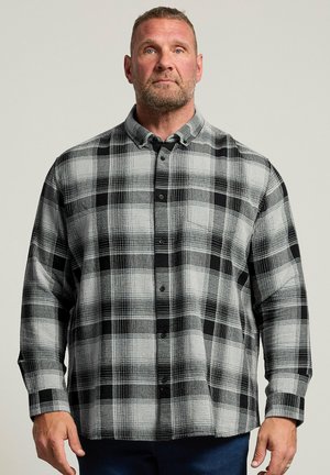 Homme avec barbe portant une chemise à carreaux noire et blanche boutonnée, debout devant un fond clair uni.