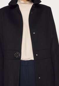 Abrigo de lana negro con cuello alto, botones a presión y un acento metálico circular en el logo. Debajo, se puede ver un suéter de cuello alto beige.