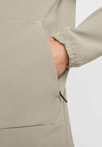 Lichtbeige jasstof met een voorzak en elastische manchetten. Gestructureerd oppervlak, minimalistisch ontwerp en een zwarte ritsdetail.