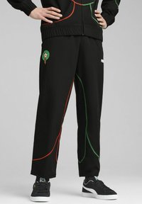 Pantalones de chándal negros con costuras en verde y rojo, cinturilla elástica y emblema marroquí en el muslo. El material parece ligero y suave.