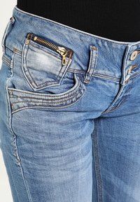 Pantalones vaqueros de mezclilla azul con un bolsillo con cremallera en la parte delantera derecha, costuras en contraste y un diseño ajustado con cintura de altura media.