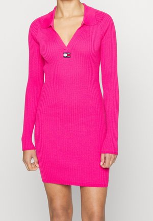 Frau trägt ein geripptes, knallpinkes, langärmeliges, knielanges Kleid mit Kragen und dem "Tommy Jeans"-Logo auf der Brust.