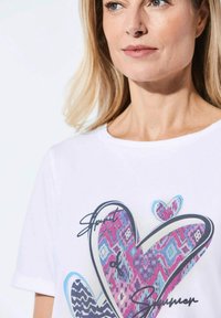 Witte t-shirt met korte mouwen, versierd met een grote hartafbeelding in kleurrijke patronen, met blauwe accenten en de tekst "Spirit of Summer" in het zwart.