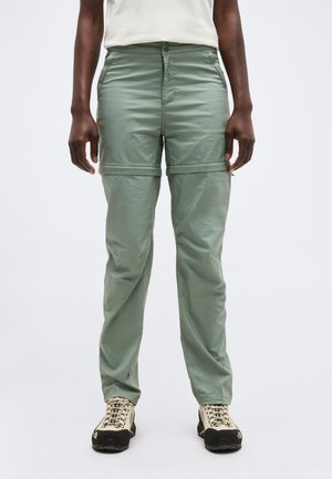 Άτομο που φοράει ανοιχτό πράσινο παντελόνι τύπου convertible με αποσπώμενα πόδια και λευκά παπούτσια πεζοπορίας, στέκεται μπροστά σε απλό φόντο.