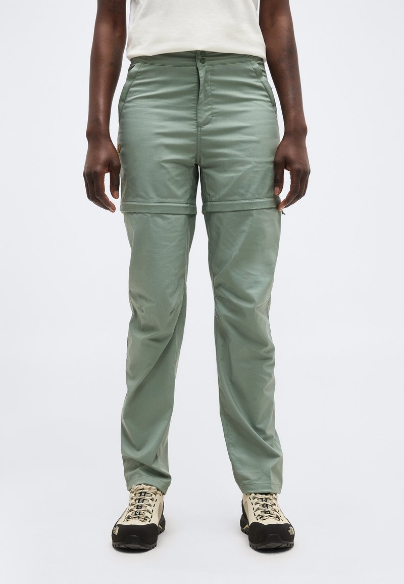 Person trägt hellgrüne wandelbare Hose mit abzipbaren Beinen und weiße Wanderschuhe, steht vor einfarbigem Hintergrund.