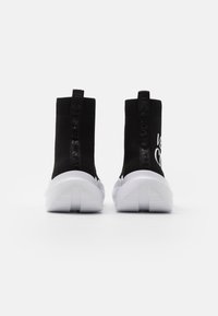 Zapatillas de punto negras con diseño de tobillo alto, suela blanca y detalles de marca texturizados en el talón. Acabado suave.