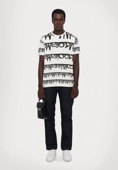 Witte graphic T-shirt met zwart tekstpatroon, gecombineerd met een donkere spijkerbroek en witte sneakers. Model houdt een kleine zwarte tas vast met een zilveren sluiting.