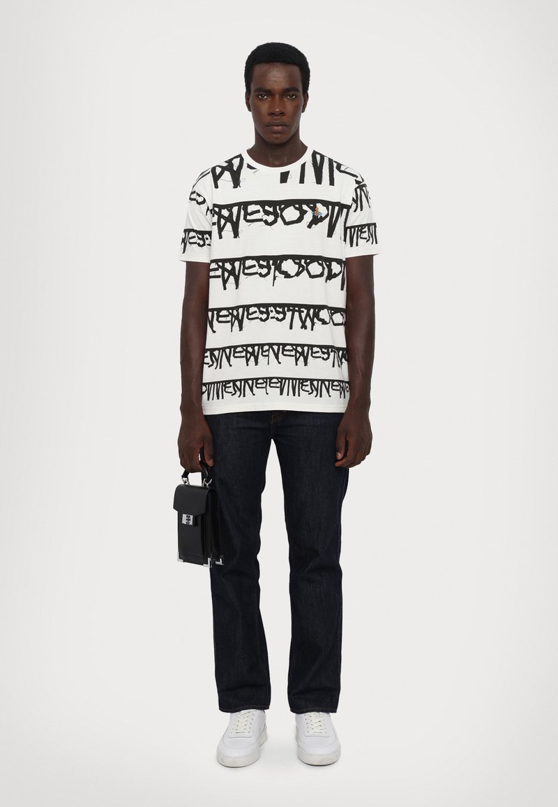 Weißes Grafik-T-Shirt mit schwarzem Textmuster, kombiniert mit dunklen Jeans und weißen Sneakers. Das Model hält eine kleine schwarze Tasche mit einem silbernen Verschluss.