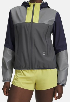 Kvinde iført en farveblocket grå, gul og marineblå lynlåshættetrøje med to lynlåslommer på brystet og gule sportsshorts.