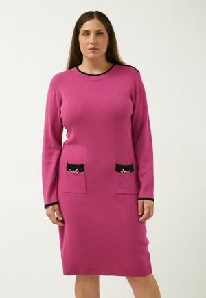 Langärmliges Kleid aus leuchtendem pinkfarbenem Stoff, mit zwei Fronttaschen mit schwarzem Besatz und goldenen Kettenakzenten. Rundhalsausschnitt und taillierte Silhouette.