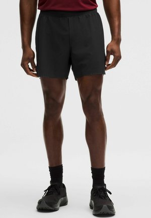 Mężczyzna w czarnych sportowych szortach, czarnych skarpetkach do kostek i czarnych butach do biegania, stojący na jasnym, jednolitym tle.