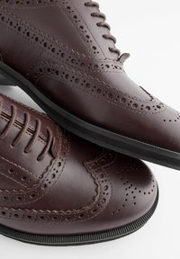 Chaussures en cuir marron avec des détails en brogue, présentant une finition lisse, un bout pointu, un design à lacets et une semelle extérieure en caoutchouc noire.