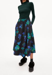 Maglietta verde a maniche lunghe con coste abbinata a una gonna nera a lunghezza ginocchio con un motivo floreale colorato e una cintura elastica. Calzature nere.