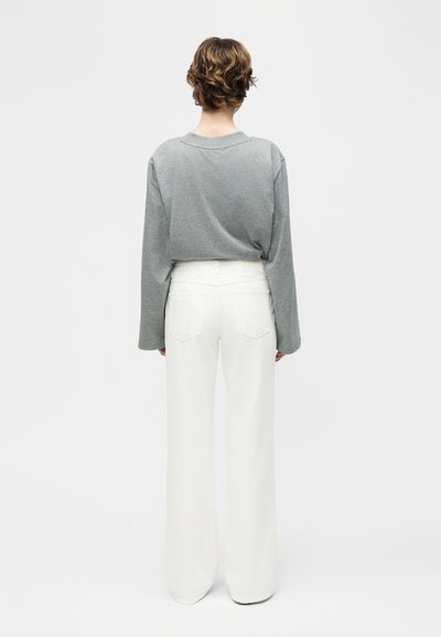 Personne aux cheveux courts et bouclés, vue de dos, portant un haut gris à manches longues et un pantalon blanc taille haute à jambes larges.