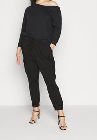 Donna che indossa un top nero a spalle scoperte con maniche lunghe, pantaloni cargo neri con cordino e sandali neri con tacco alto su sfondo bianco.