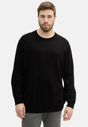 Black long-sleeve sweater with a ribbed texture, round neckline, and a small logo detail on the chest, worn with gray pants.