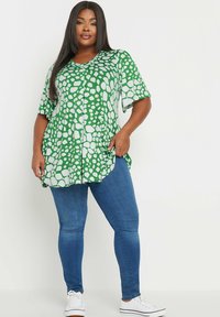 Grön kortärmad topp med ett vitt abstrakt mönster, tillsammans med blå smala jeans och vita sneakers. Toppens design har en V-ringning och avslappnad passform.