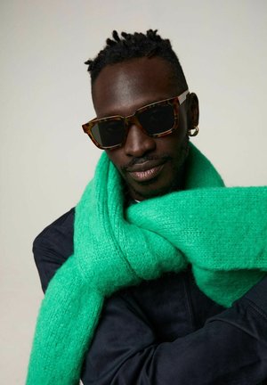 Schildpadbril, gestructureerde groene sjaal gemaakt van zacht garen en een donkere jas. De sjaal is om de nek gewikkeld, wat contrast biedt.