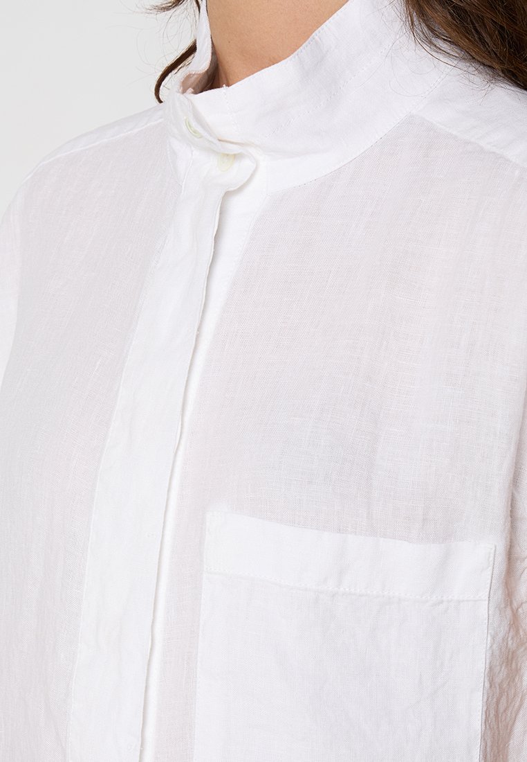 Weißes Leinenhemd mit hochgeschlossenem Kragen, mittigen Knöpfen und einer Brusttasche auf der linken Seite. Strukturierter Stoff mit leicht zerknittertem Aussehen.