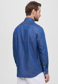Camicia in denim blu con colletto rialzato, chiusura frontale a bottoni, maniche lunghe e orlo curvo. Tessuto liscio e design classico.