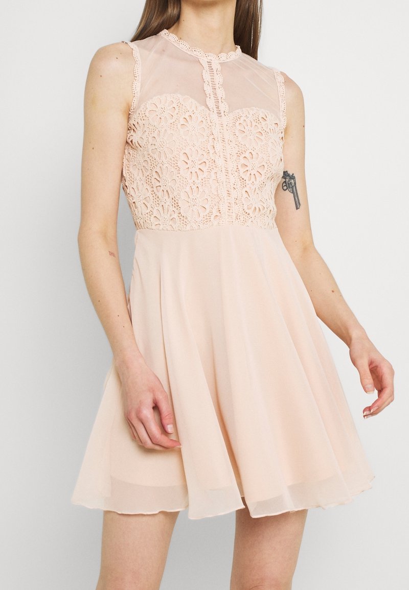 Robe de couleur pêche avec un corsage en dentelle, un décolleté transparent, des détails de boutons et une jupe en chiffon évasée, présentant une bordure en festons.