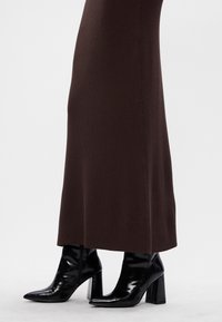 Lśniące czarne botki na obcasie z ostrym noskiem, noszone pod długą, ciemnobrązową, prążkowaną spódnicą.