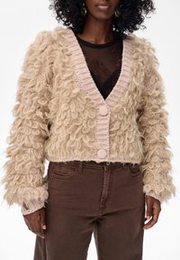 Czarna kobieta w beżowym kardiganie z pętli włóczki, nosząca czarną przezroczystą bluzkę w kwiaty i brązowe spodnie z wysokim stanem, stojąca na białym tle.