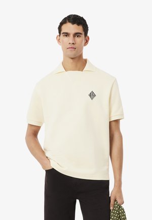 Jonge man draagt een crèmekleurig poloshirt met korte mouwen en een zwart diamantenlogo, zwarte broek, en houdt een patroonstof in zijn rechterhand.