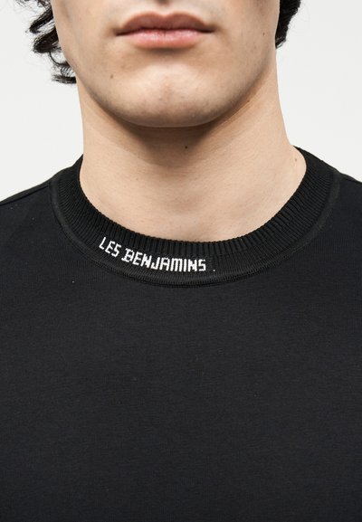 Κοντινό πλάνο του κάτω μέρους του προσώπου και του λαιμού ενός ατόμου που φοράει μαύρο πουκάμισο με το κέντημα "LES BENJAMINS" σε λευκό χρώμα στο γιακά.