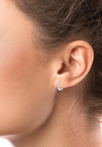 Srebrni uhani v obliki polmeseca s majhnimi vdelanimi prozornimi kamni, prikazani na ušesu proti nevtralni ozadju.