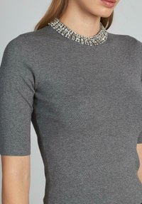 Top grigio a maniche corte con colletto gioiello decorato con elementi metallici e trasparenti, creando un collo testurizzato e decorativo.