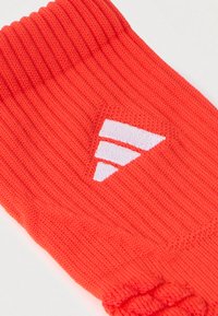 Rød strikket sok med ribbet top, med et hvidt logodesign bestående af tre parallelle striber. Blød tekstur, standardstørrelse.