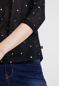 Svart topp med trekvartsärmar och silverprickiga mönster, tillverkad av ett texturerat tyg. Inkluderar en liten sidetikett.