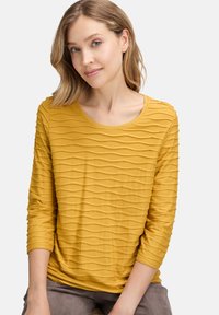 Senfgelbes Langarm-T-Shirt mit einem strukturierten wellenförmigen Muster, runder Ausschnitt und 3/4-Ärmeln, aus einem weichen Stoff hergestellt.