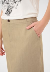 Un pantalon beige à la texture lisse, doté d'une fermeture à bouton, de poches latérales et d'une coupe ajustée, porté avec un haut blanc à motif.