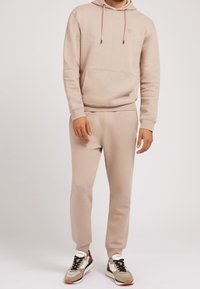 Sudadera beige y joggers hechos de una tela suave, con un bolsillo canguro, capucha con cordón y piernas ajustadas. Combinados con zapatillas deportivas.
