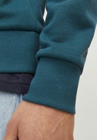 Sudadera verde azulado con puños acanalados. La tela es suave con una textura lisa. Los puños contrastan con un dobladillo inferior de un azul marino oscuro.