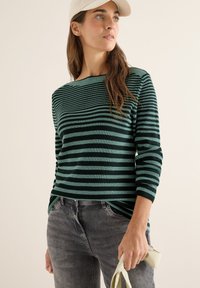 Maglione lavorato a maglia a righe verdi con scollo a barca, maniche lunghe e vestibilità comoda, abbinato a jeans grigi e con una borsa di colore chiaro.