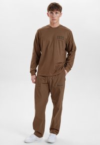 Camicia marrone a maniche lunghe con "1935" stampato sul petto, abbinata a pantaloni cargo marroni con tasche e un logo blu, indossata con sneaker bianche.