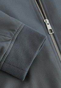 Manche de veste gris foncé avec poignet côtelé à côté d’une fermeture éclair métallique argentée partiellement zippée sur tissu côtelé.