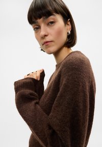 Καφέ, υφασμάτινος πουλόβερ με υφή, με μακριά μανίκια, ριμπ μανσέτες και στρογγυλή λαιμόκοψη. Το μαλακό ύφασμα φαίνεται άνετο και ζεστό. Το μοντέλο έχει ουδέτερο στυλ.