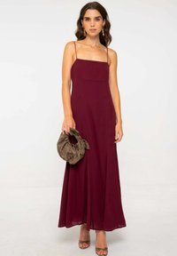 Robe maxi bourgogne avec des bretelles fines, un décolleté carré et une jupe fluide. Le mannequin tient un sac en tissu à motifs. Des talons complètent le look.