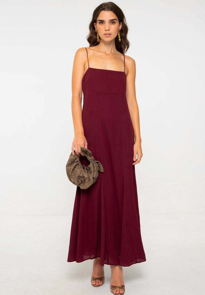 Robe maxi bourgogne avec des bretelles fines, un décolleté carré et une jupe fluide. Le mannequin tient un sac en tissu à motifs. Des talons complètent le look.