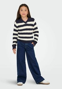 Chica con cabello largo y oscuro, vestida con un suéter de rayas azul marino y blancas, pantalones vaqueros anchos de un azul oscuro, zapatillas blancas, de pie con una mano en el bolsillo.