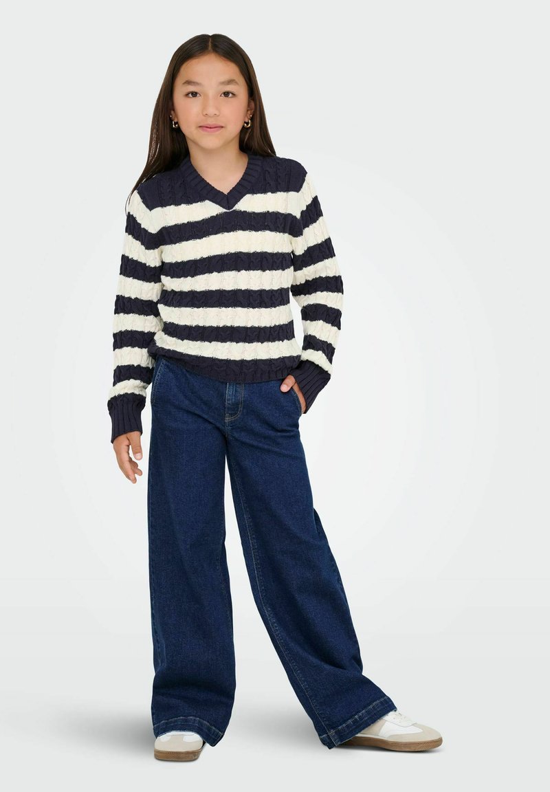 Chica con cabello largo y oscuro, vestida con un suéter de rayas azul marino y blancas, pantalones vaqueros anchos de un azul oscuro, zapatillas blancas, de pie con una mano en el bolsillo.