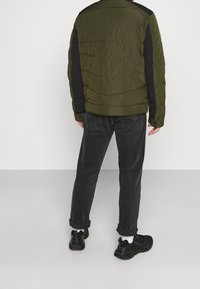 Dunjakke i olivengrøn med sorte accenter, der har en tekstureret overflade og en afslappet pasform. Båret med mørke jeans og sorte sneakers.