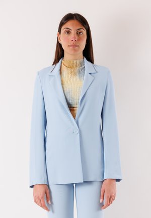 Donna con capelli lunghi e scuri che indossa una giacca a vento azzurra chiara sopra una maglia a collo alto con trama, in piedi davanti a uno sfondo bianco semplice.