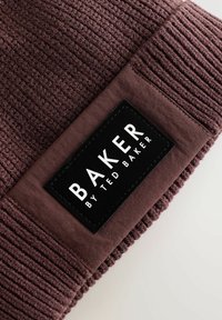 Gorro de punto burdeos con una textura acanalada y una etiqueta negra plana con el texto blanco "BAKER by TED BAKER" en el lateral.